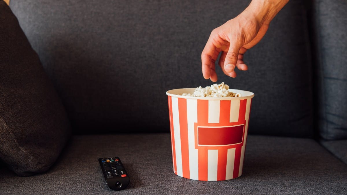 Cropped view of man reaching popcorn in bucket near remote