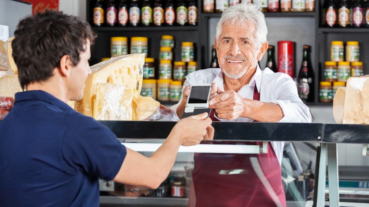 Salesman Accepting Mobile Payment From Customer In Cheese Shop